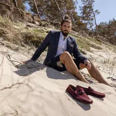 Mężczyzna w marynarce i szortach siedzi na plaży. Obok niego leżą bordowe mokasyny, idealne jako buty do cygaretek.