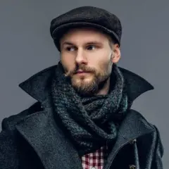 Mężczyzna w stylowym, wełnianym płaszczu i szaliku, z charakterystycznym wąsem i brodą, nosi ciemne nakrycie głowy.