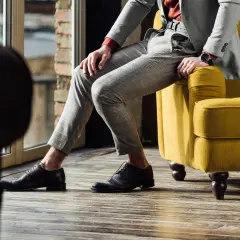 Eleganckie, brązowe oksfordy to idealne buty do szarego garnituru w kratę. Mężczyzna siedzi na żółtej kanapie.