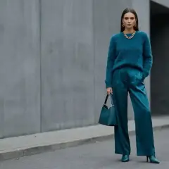 Elegancka kobieta w stylizacji w kolorze petrol, z błyszczącymi spodniami i miękkim swetrem, pozuje na tle betonowej ściany.