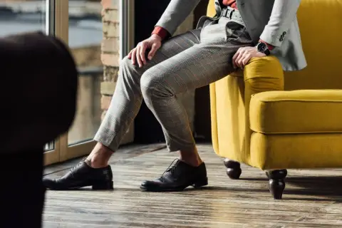 Eleganckie, brązowe oksfordy to idealne buty do szarego garnituru w kratę. Mężczyzna siedzi na żółtej kanapie.