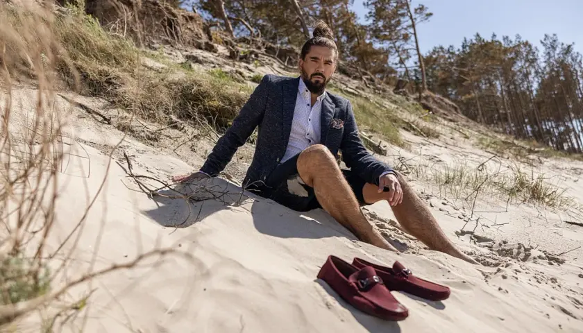 Mężczyzna w marynarce i szortach siedzi na plaży. Obok niego leżą bordowe mokasyny, idealne jako buty do cygaretek.