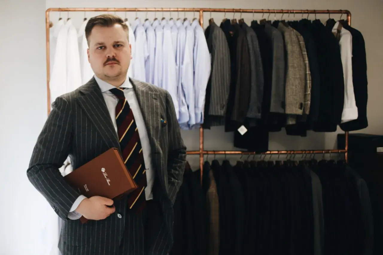 Elegancki mężczyzna w garniturze w prążki trzyma teczkę. Za nim wiszą marynarki i koszule.
