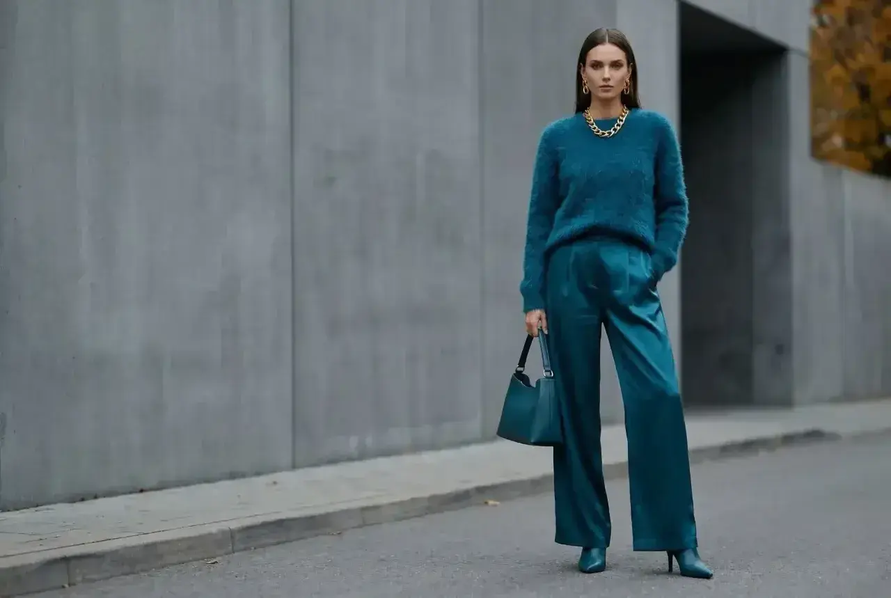 Elegancka kobieta w stylizacji w kolorze petrol, z błyszczącymi spodniami i miękkim swetrem, pozuje na tle betonowej ściany.
