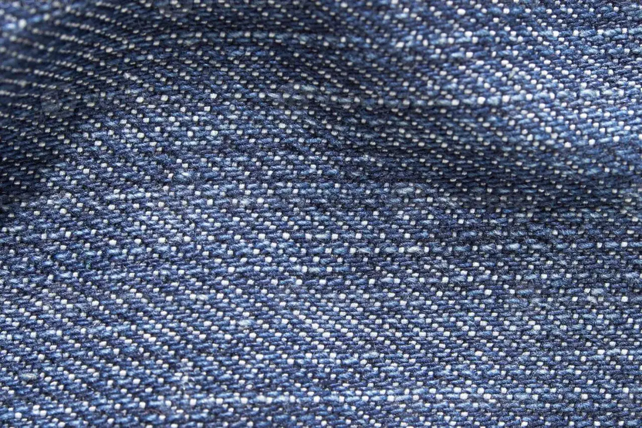 Zbliżenie na niebieski materiał, który jest przykładem tego, co to jest denim. Widać charakterystyczny, skośny splot i drobne białe nitki.