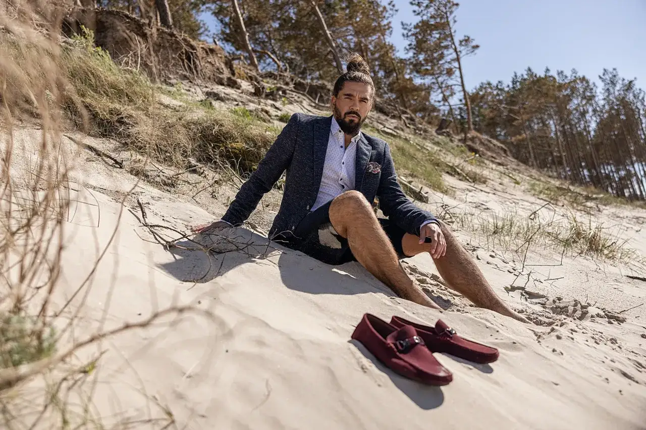 Mężczyzna w marynarce i szortach siedzi na plaży. Obok niego leżą bordowe mokasyny, idealne jako buty do cygaretek.